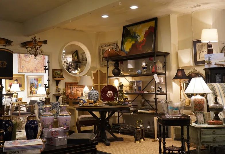 A corner of the M&eacute;lange showroom: a large round mirror, a black iron &eacute;tag&egrave;re loaded with accessories, table lamps, blue-and-gold porcelain vases and a wall of framed art above a console.