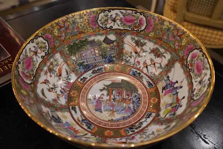 A Chinese Rose Medallion porcelain bowl with a gilt rim, hand-painted inside with court figures, birds, peonies and foliage, standing on a dark surface beside books.