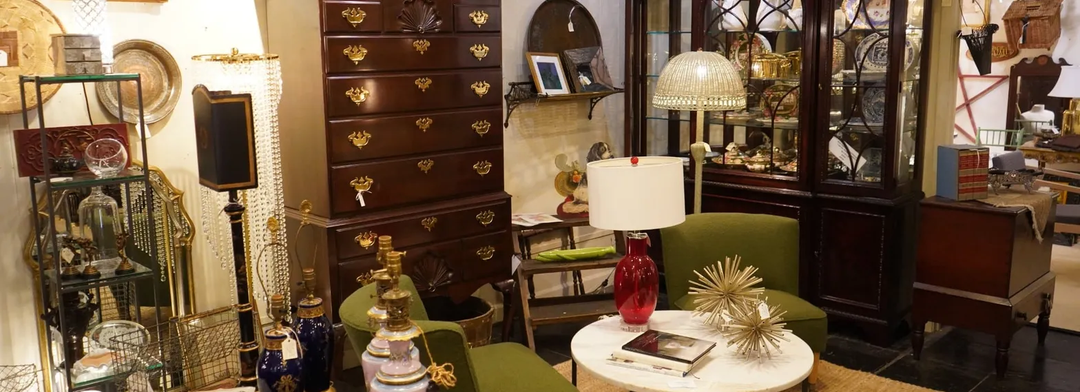 A styled room inside M&eacute;lange: a mahogany chest-on-chest with a broken-arch pediment, a glazed mahogany breakfront of china behind, two olive-green upholstered chairs around a round white marble-topped table, a red glass lamp and a gilt starburst on the table.