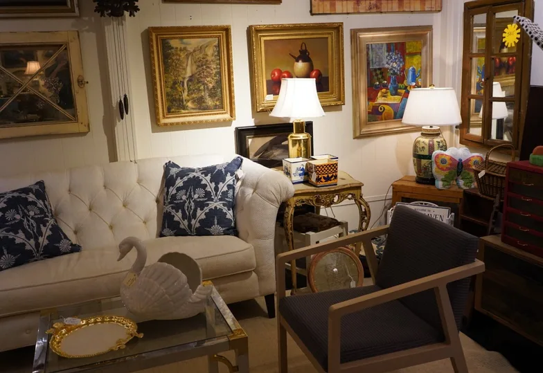 A cream tufted sofa with blue-and-white patterned pillows, a glass swan and a gilt tray on a glass coffee table, a mid-century wood-arm chair, and a wall hung edge to edge with framed paintings and mirrors.