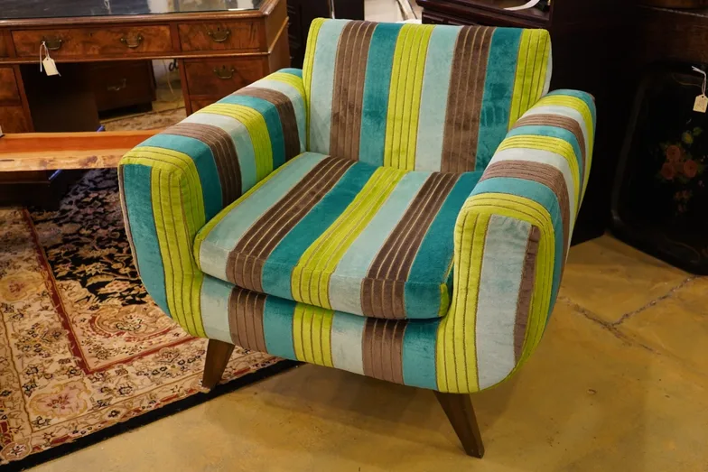 A striped velvet armchair in turquoise, chartreuse and taupe on splayed wooden legs, standing on a patterned rug in front of a burl-wood desk.
